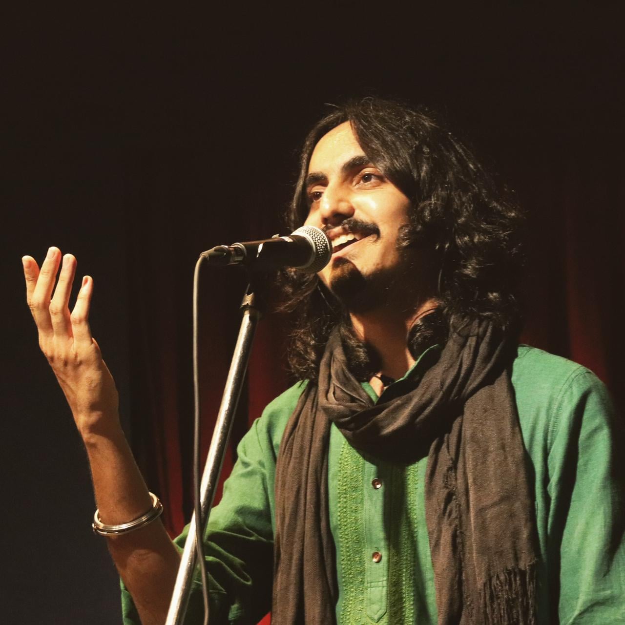 Aditya Gadhvi singing into a microphone on stage, one hand raised, in a green kurta and dark scarf